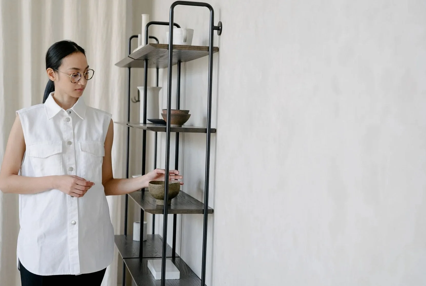 A woman in a white shirt arranges minimalist home decor on an industrial-style shelving unit.